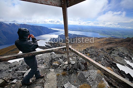 Tramper / Hiker photographing on top of Mount Luxmore trig (1472m) on Kepler Track Great Walk. South Fiord of Lake Te Anau at left, Fiordland National Park, Southland District, Southland Region, New Zealand (NZ)