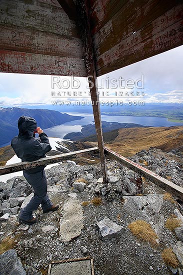 Tramper / Hiker with camera on top of Mount Luxmore trig (1472m) on Kepler Track Great Walk. South Fiord of Lake Te Anau at left, Fiordland National Park, Southland District, Southland Region, New Zealand (NZ)